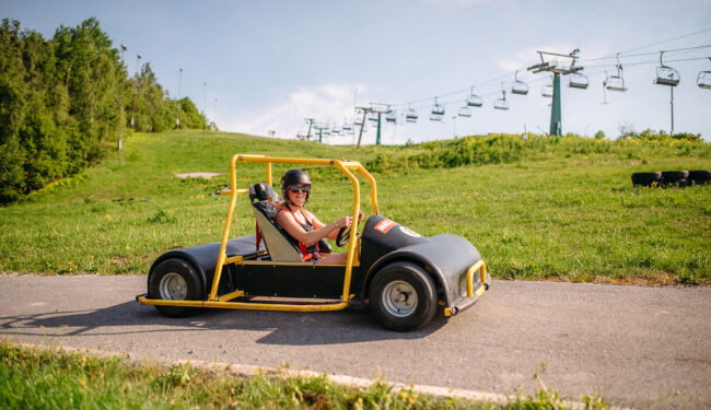 Bilden visar Flottsbro Downhill Cars, aktivitet med gokart sedd från sidan kör på en väg i en backe med grönt gräs och blå himmel bakom.