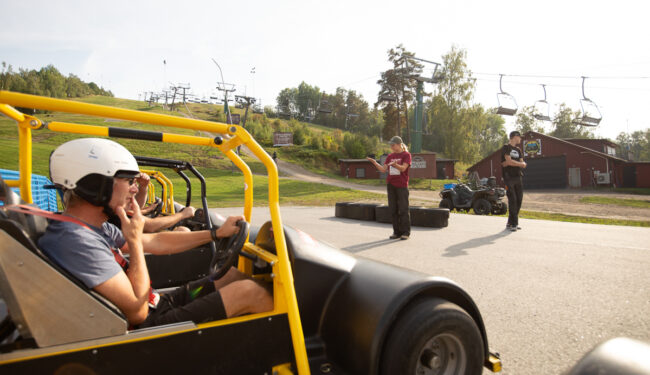 Bild med en gokart och förare från sidan frmför och längre bort i bild står en person som ger instruktioner.