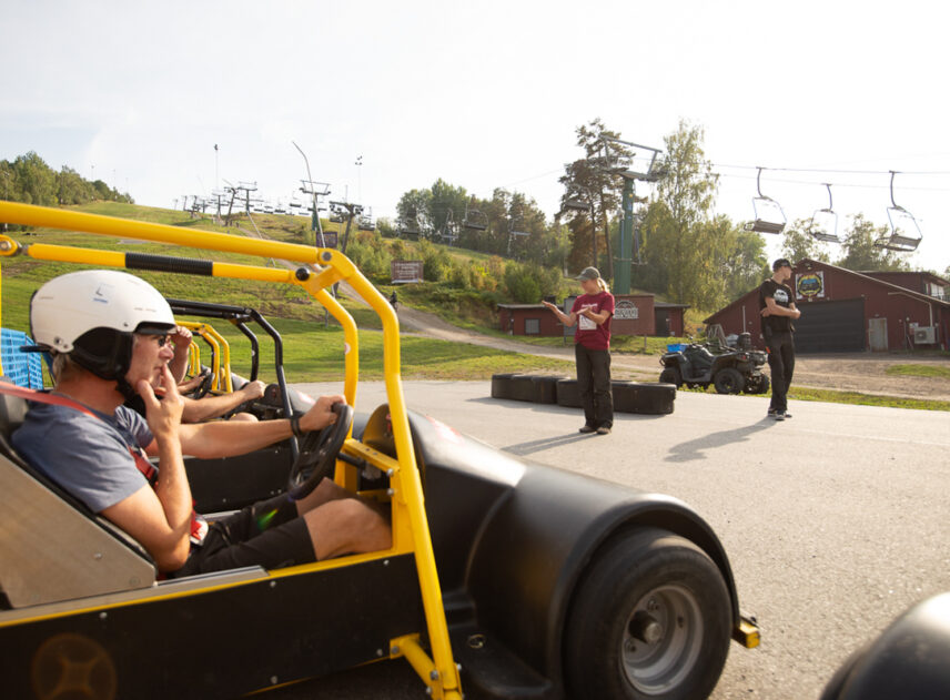 Bild med en gokart och förare från sidan frmför och längre bort i bild står en person som ger instruktioner.