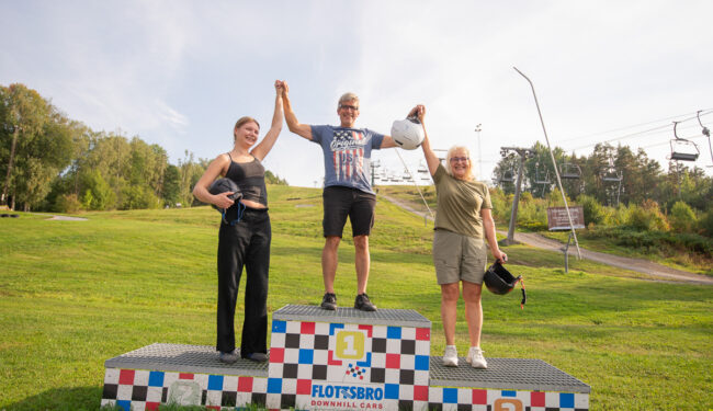 3 personer på en prispall, de håller händerna uppsträckta i luften på prispallen för Downhill Cars