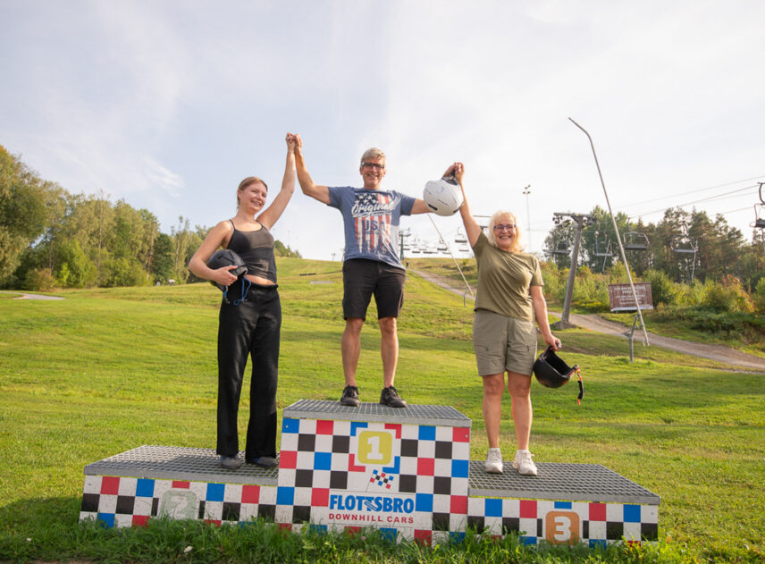 3 personer på en prispall, de håller händerna uppsträckta i luften på prispallen för Downhill Cars