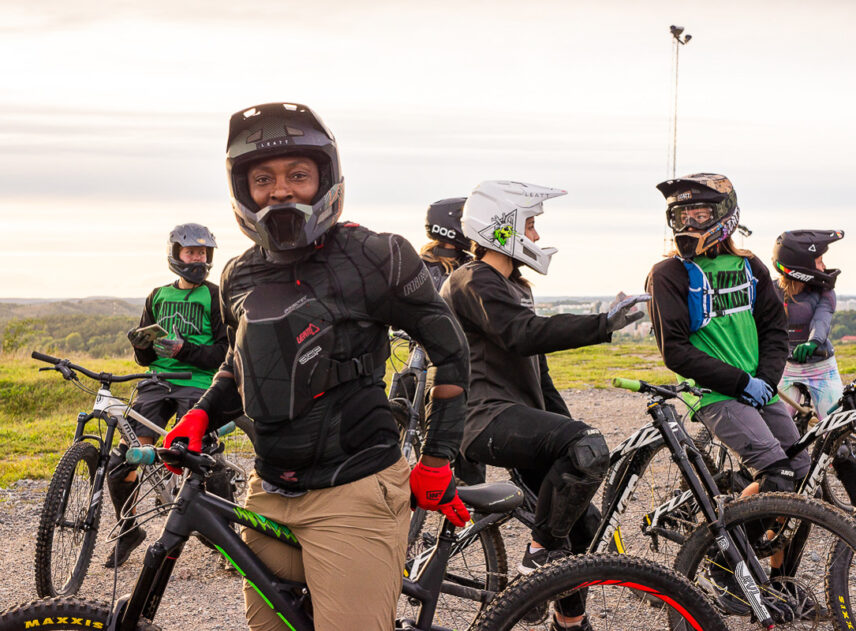 En grupp tjejer med cyklar iförda cykelutrustning med hjälmar i förgrunden ser den närmaste cyklisten in i kameran