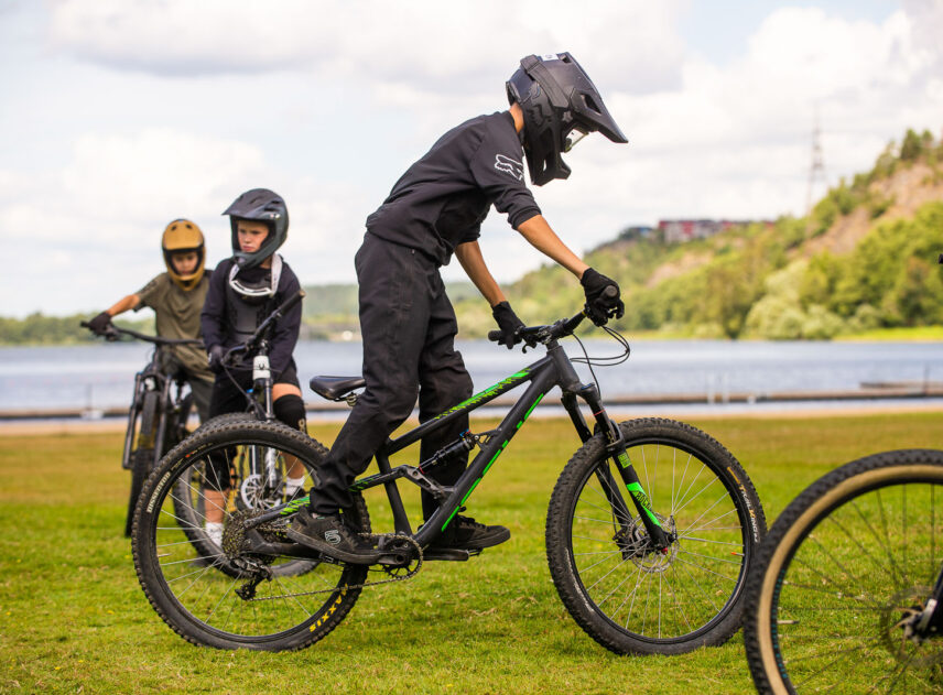 Tre ungdomar tränar balans på cykel, i bakgrunden syns en sjö.