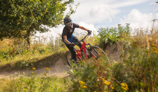Ung cyklist på downhill bana mot blå himmel, i förgrunden syns blommor.