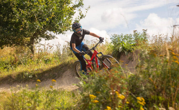 Ung cyklist på downhill bana mot blå himmel, i förgrunden syns blommor.