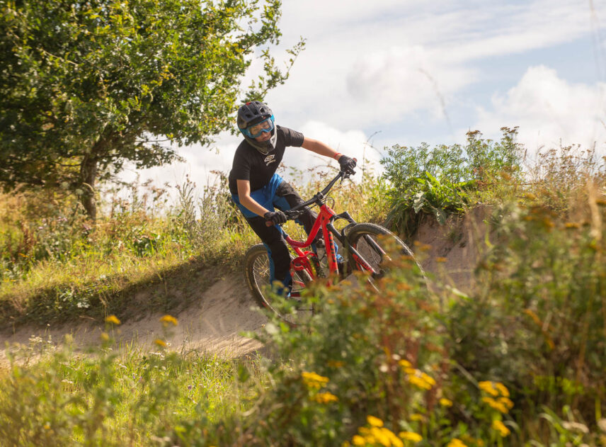 Ung cyklist på downhill bana mot blå himmel, i förgrunden syns blommor.