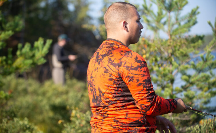 Två personer som står i tät vegetation och fiskar, i närbild syns en man i orange tröja och längre bort skymtas en annan person hållande fiskespö.