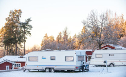 Vinterbild på campingplats, två husvagnar är täckta med snö, i bakgrunden skymtar röda stugor.