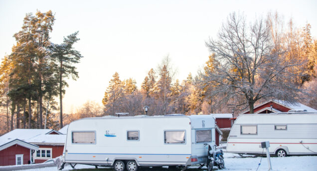 Vinterbild på campingplats, två husvagnar är täckta med snö, i bakgrunden skymtar röda stugor.