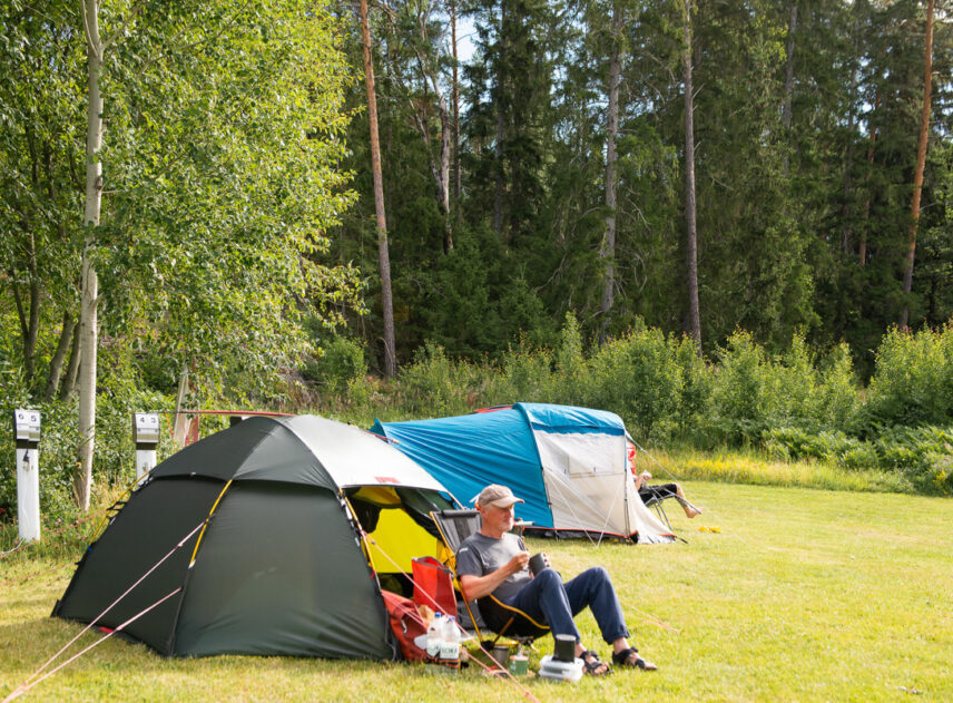 Bild på en man som sitter utanför ett grönt tält på en campingplats, i bakgrunden syns en skog.