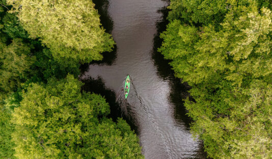 Bild tagen ovanifrån på en kanot i en kanal, runt omring syns skog och vattnet är mörkt.