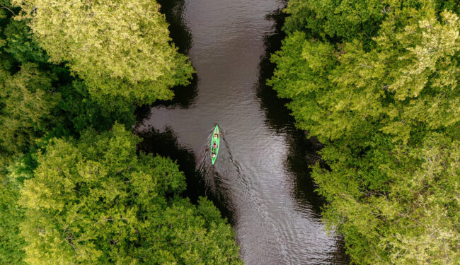 Bild tagen ovanifrån på en kanot i en kanal, runt omring syns skog och vattnet är mörkt.