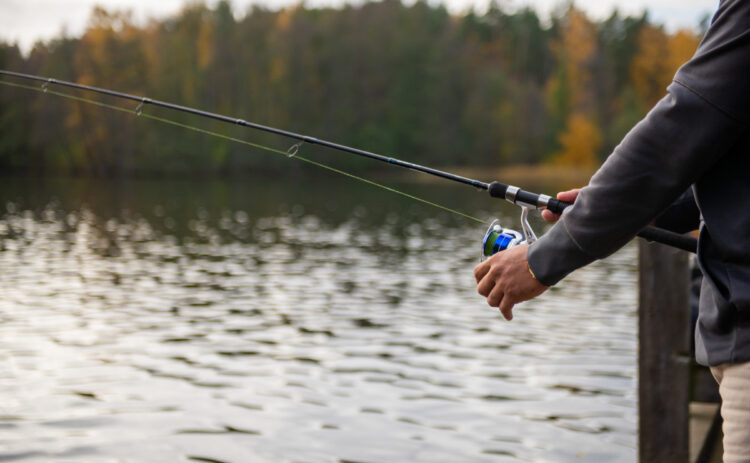 En bild på en beskuren person som fiskar med kastspö vid en sjö omgiven av skog.