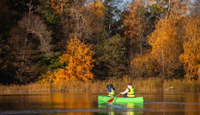 Bilden visar en kanot med 2 personer som paddlar i Albysjön en höstdag, i bakrunden syns lövträd i skiftande höstfärger.