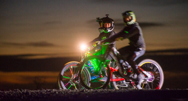 Bild tagen på kvällen på Flottsbro Nightride, cyklister med ljus centralt i bilden, det är mörk bakgrund.