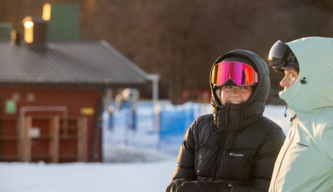 Två skidåkare som talar med varandra sedda i profil, i bakgrunden syns anläggningens lifthus.