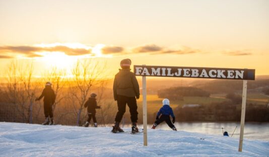 Bild från toppen av Flottsbro skidbacke i solnedgången, skidåkare syns sedda bakifrån, i förgrundens syns en skylt med texten familjebacken.