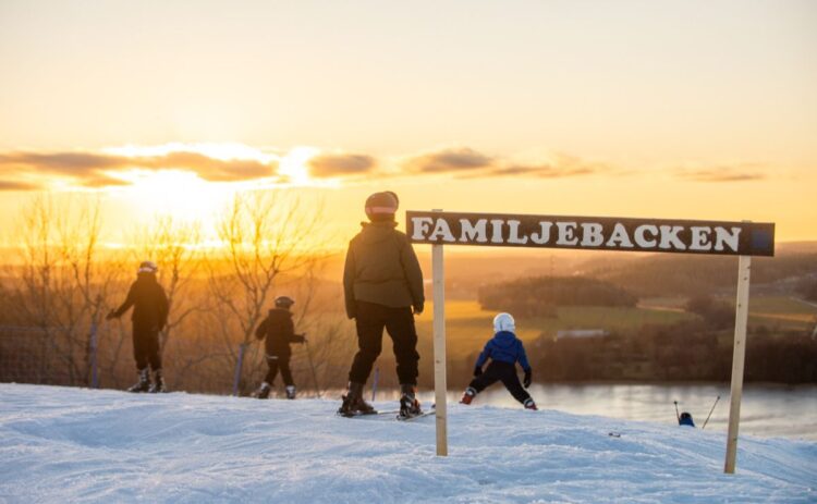 Bild från toppen av Flottsbro skidbacke i solnedgången, skidåkare syns sedda bakifrån, i förgrundens syns en skylt med texten familjebacken.