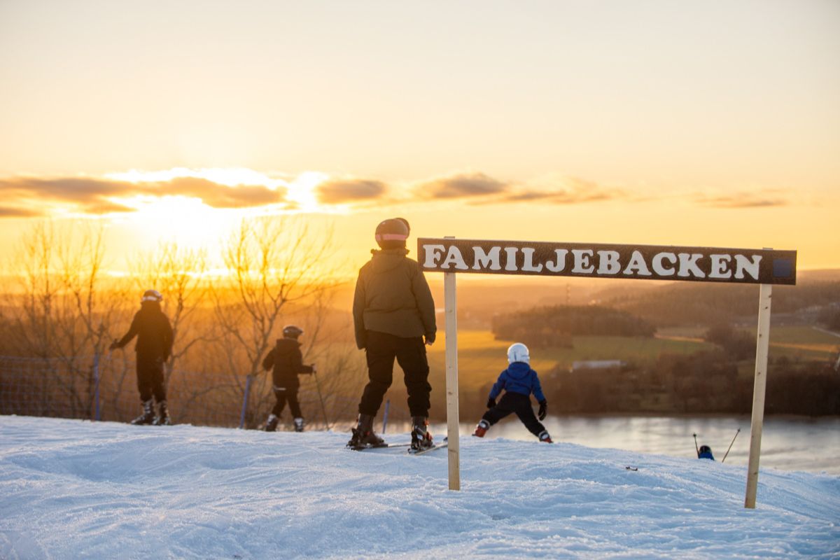 Bild från toppen av Flottsbro skidbacke i solnedgången, skidåkare syns sedda bakifrån, i förgrundens syns en skylt med texten familjebacken.