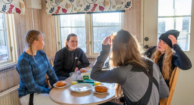 Fyra tjejer sitter runt ett bord iförda skidutrustning i en liten stuga.