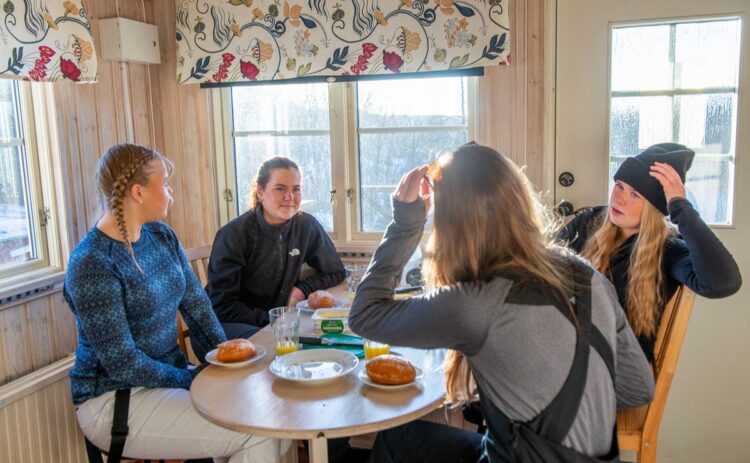 Fyra tjejer sitter runt ett bord iförda skidutrustning i en liten stuga.
