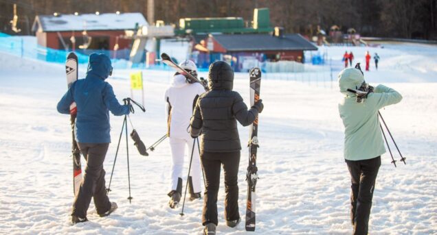 Fyra personer sedda bakifrån går mot en skidlift, samtliga bär på skidutrustning.