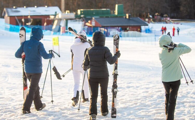 Fyra personer sedda bakifrån går mot en skidlift, samtliga bär på skidutrustning.