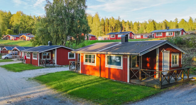 Bilden visar röda en grusväg med röda sommarstugor som ligger på rad, gräsmattor är göra och himlen är blå.