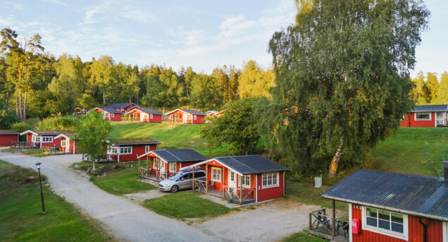 Bilden visar röda en grusväg med röda sommarstugor som ligger på rad, gräsmattor är göra och himlen är blå.