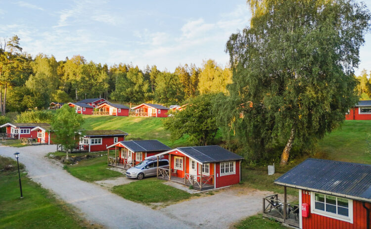 Bilden visar röda en grusväg med röda sommarstugor som ligger på rad, gräsmattor är göra och himlen är blå.