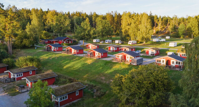 Bilden visar rรถda en grusvรคg med rรถda sommarstugor som ligger pรฅ rad, grรคsmattor รคr gรถra och himlen รคr blรฅ.