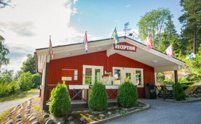 Röd stuga med skylten reception ovanför dörren, vid taknocken hänger olika flaggor och framför stugan står 3 buskar.