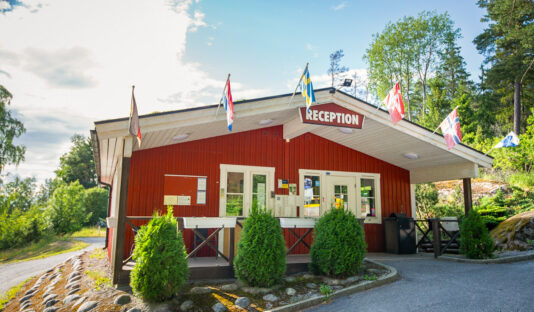 Röd stuga med skylten reception ovanför dörren, vid taknocken hänger olika flaggor och framför stugan står 3 buskar.