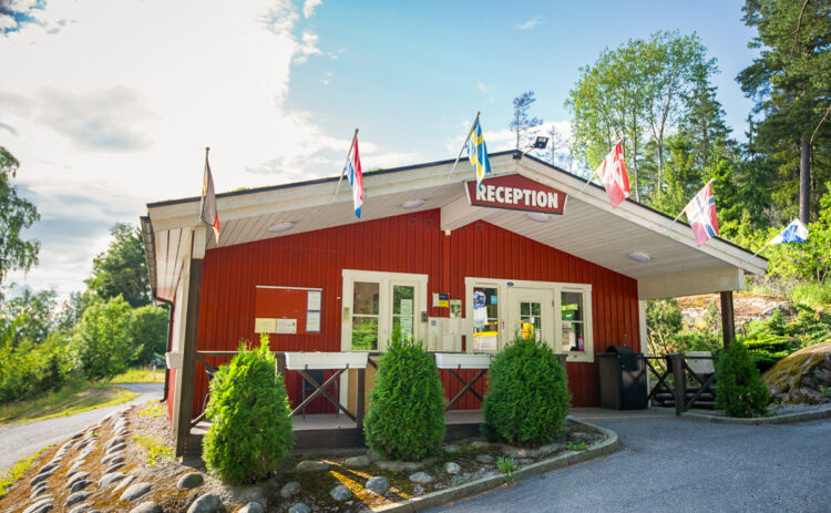 Röd stuga med skylten reception ovanför dörren, vid taknocken hänger olika flaggor och framför stugan står 3 buskar.