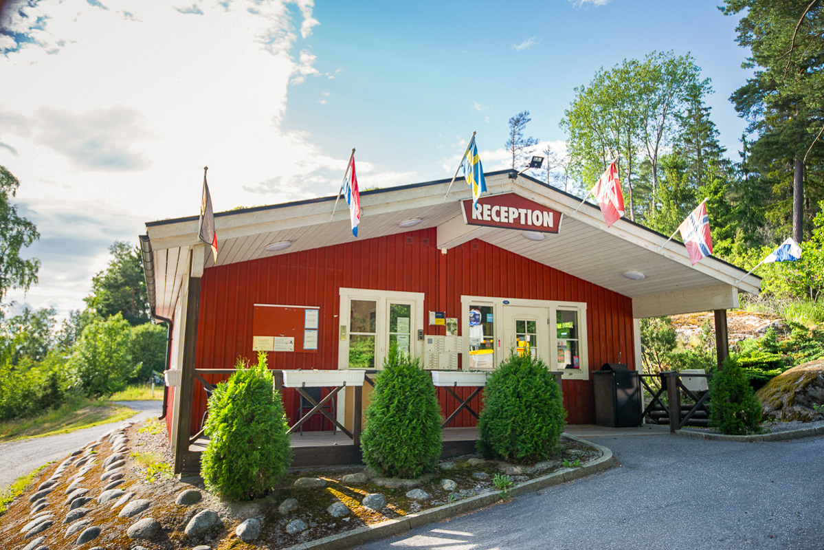 Röd stuga med skylten reception ovanför dörren, vid taknocken hänger olika flaggor och framför stugan står 3 buskar.