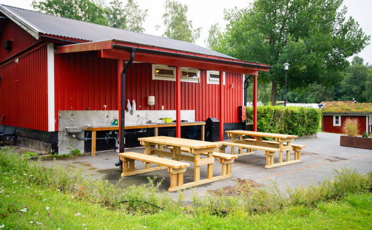 Röd servicebyggnad sedd från sidan, utmed byggnaden finns ett utomhuskök och framför står det 2 matbord.
