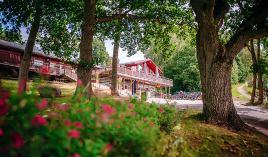 Röd lång byggnad skymtas mellan träden, i förgrunden syns rosa blommor.