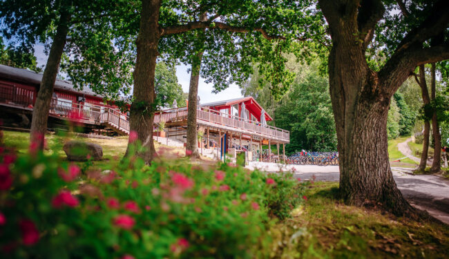 Röd lång byggnad skymtas mellan träden, i förgrunden syns rosa blommor.