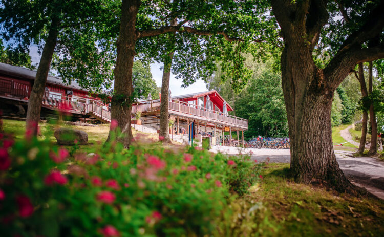 Röd lång byggnad skymtas mellan träden, i förgrunden syns rosa blommor.