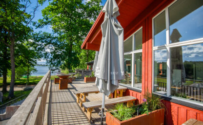 Bild på terass framför röd byggnad en solig sommardag, i bakgrunden syns grönskande lövträd och blå himmel.