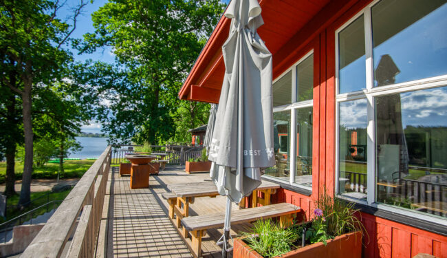 Bild på terass framför röd byggnad en solig sommardag, i bakgrunden syns grönskande lövträd och blå himmel.