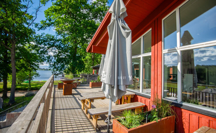 Bild på terass framför röd byggnad en solig sommardag, i bakgrunden syns grönskande lövträd och blå himmel.