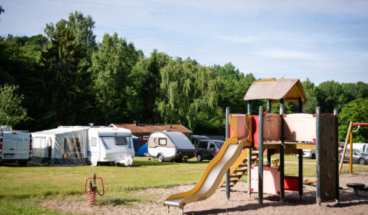 En lekplats en solig sommardag, i bakgrunden syns husvagnar och grön skog, himlen är blå.