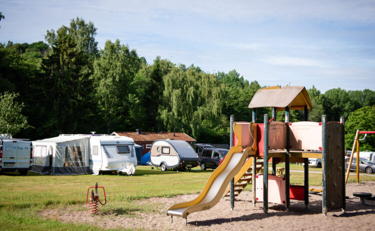 Bild på Flottsbro Camping Stockholm som visar en lekplats en solig sommardag, i bakgrunden syns husvagnar och grön skog, himlen är blå.