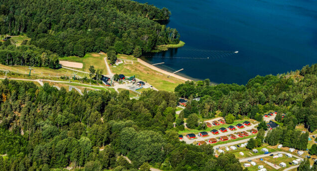 Ett flygfoto på område som visar flottbros backe på sommaren, uppe till höger i bild syns sjön och nedtill syns ett campingområde.