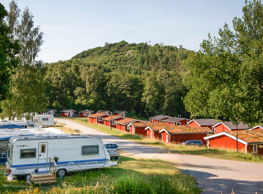 Bild på campingplats som med husvagn parkerad framför en rad röda stugor, gräsmattan är grön och i bakgrunden syns en grönskande kulle.