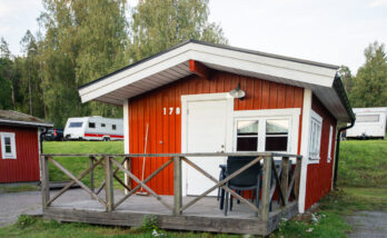 Bild på röd stuga sedd rakt framifrån, i bakgrunden syns två husvagnar.