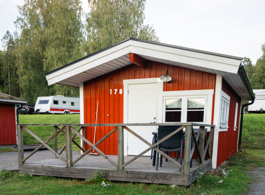 Bild på röd stuga sedd rakt framifrån, i bakgrunden syns två husvagnar.