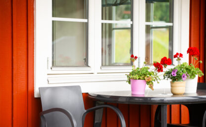 Bild på fönstret på en röd stuga, väggen är röd och framför fönstret står ett bord med blommor på.