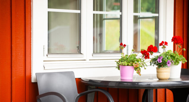 Bild på fönstret på en röd stuga, väggen är röd och framför fönstret står ett bord med blommor på.